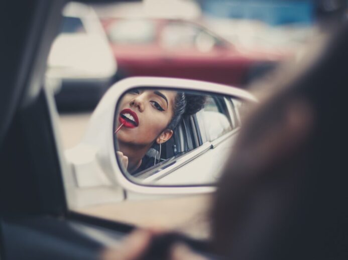 woman putting liquid lipstick on her lips while looking at vehicle's mirror during daytime