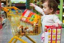 toddle carrying red and white box standing beside yellow shopping cart