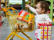 toddle carrying red and white box standing beside yellow shopping cart
