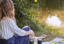 woman in white shirt and blue denim jeans sitting on green grass near lake during daytime