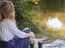 woman in white shirt and blue denim jeans sitting on green grass near lake during daytime