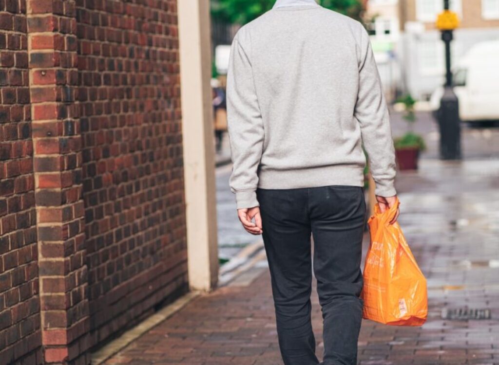man in gray hoodie and black pants holding orange plastic bag walking on sidewalk during daytime