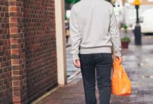 man in gray hoodie and black pants holding orange plastic bag walking on sidewalk during daytime
