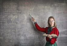 Serious female teacher wearing old fashioned dress and eyeglasses standing with book while pointing at chalkboard with schemes and looking at camera