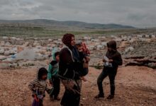 Poor ethnic woman with toddler in arms and kids near settlement surrounded with mountains under cloudy gloomy sky
