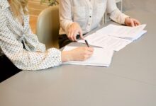 woman signing on white printer paper beside woman about to touch the documents