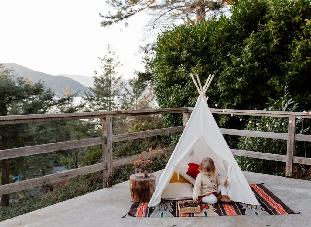 Adorable little kid sitting on rug in wigwam located on terrace with wooden border and scenic view