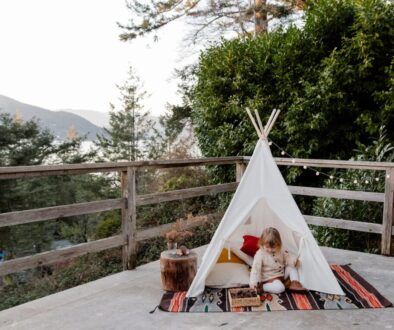 Adorable little kid sitting on rug in wigwam located on terrace with wooden border and scenic view