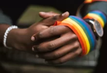 Crop anonymous African American homosexual male wearing colorful rainbow ribbon on arm while sitting on street with hands clasped