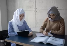 Concentrated multiracial female students in headscarves taking notes in workbooks while sitting at table with textbook during lesson preparation in university