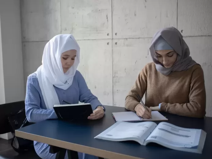 Concentrated multiracial female students in headscarves taking notes in workbooks while sitting at table with textbook during lesson preparation in university