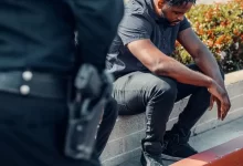 Man in Gray Button Up Shirt and Gray Denim Jeans Sitting on Concrete Block Beside a Police Officer