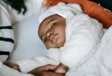 From above of crop anonymous African American mother holding hand of cute newborn baby in warm clothes sleeping on cozy bed