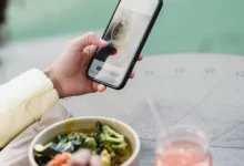 Crop unrecognizable female blogger touching screen on cellphone above street cafe table with healthy food and smoothie