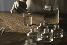 Crop anonymous female in latex gloves making fluid for perfume among glass jars on wooden table