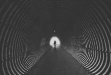 grayscale photo of man holding his bicycle in tunnel