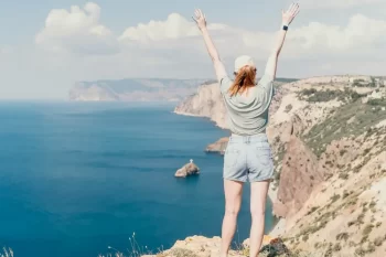Žena na vrchu útesu zdvihá ruce vzhůru.