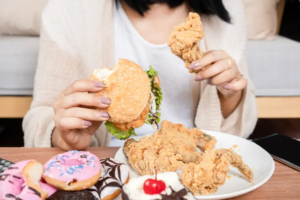 shutterstock_2257686005 - Pravda24 Žena jí burger se stripsy a na stole má koblihy a buchty.