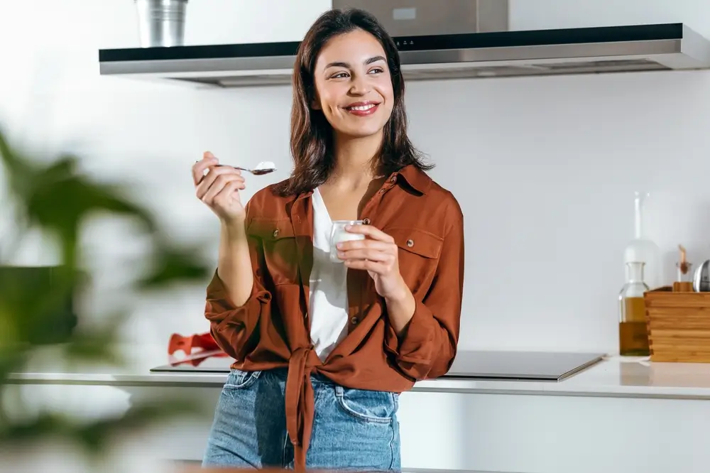 shutterstock_2339463527 - Pravda24 Žena stojí v kuchyni a jí jogurt.