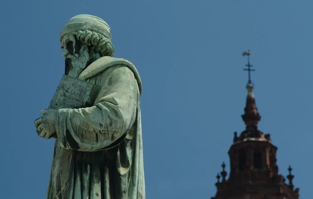 - Pravda24 Socha Johannese Gutenberga držící knihu, v pozadí věž kostela pod modrým nebem