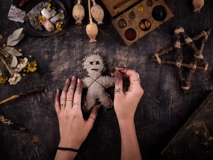 Ruce držící ručně vyrobenou voodoo panenku s jehlami, obklopenou přírodními artefakty, sušenými květy a dřevěnými nástroji při rituálu