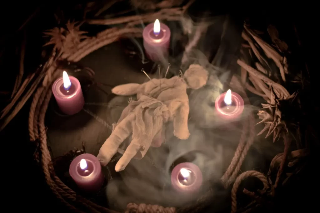 Rituální scéna s látkovou voodoo panenkou s červeným srdcem a černými vlasy, položenou na papírový pentagram, obklopenou svíčkami, jehlami a magickými předměty