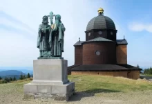 Bronzová socha věrozvěstů Cyrila a Metoděje s křížem a knihou před dřevěnou kaplí svatého Cyrila a Metoděje na Radhošti v Beskydech