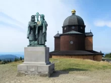 Bronzová socha věrozvěstů Cyrila a Metoděje s křížem a knihou před dřevěnou kaplí svatého Cyrila a Metoděje na Radhošti v Beskydech