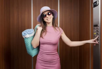 Žena v růžových šatech, klobouku a slunečních brýlích mačká tlačítko výtahu