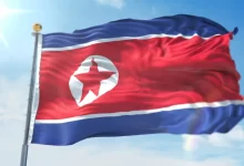Vlající státní vlajka Severní Koreje s červenou hvězdou a modrými pruhy proti modré obloze
