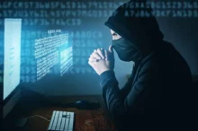 Muž v kapuci a roušce sedí před počítačem s digitálním kódem na obrazovce, působí jako hacker
