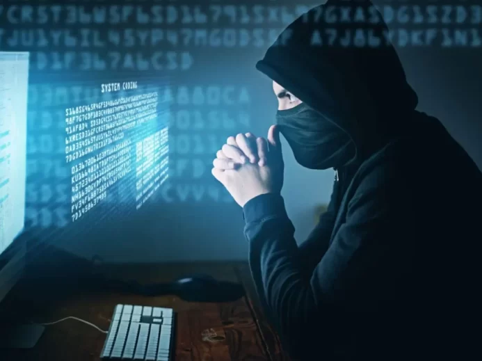 Muž v kapuci a roušce sedí před počítačem s digitálním kódem na obrazovce, působí jako hacker