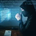 Muž v kapuci a roušce sedí před počítačem s digitálním kódem na obrazovce, působí jako hacker