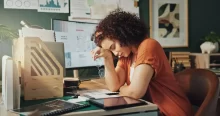 Mladá žena sedí u pracovního stolu, opřená unaveně o ruku, působí vyčerpaně a je obklopena pracovními materiály