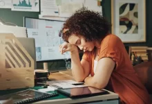 Mladá žena sedí u pracovního stolu, opřená unaveně o ruku, působí vyčerpaně a je obklopena pracovními materiály