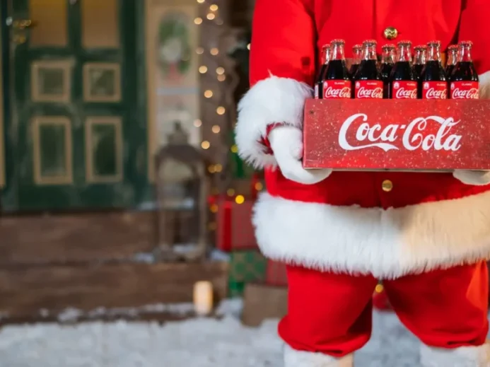Postava Santa Clause v červeném obleku s bílým lemem drží bednu se skleněnými lahvemi Coca Coly před vánočně ozdobeným domem