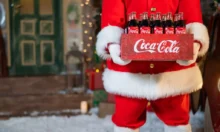 Postava Santa Clause v červeném obleku s bílým lemem drží bednu se skleněnými lahvemi Coca Coly před vánočně ozdobeným domem