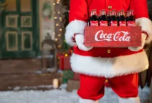 Postava Santa Clause v červeném obleku s bílým lemem drží bednu se skleněnými lahvemi Coca Coly před vánočně ozdobeným domem