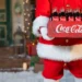 Postava Santa Clause v červeném obleku s bílým lemem drží bednu se skleněnými lahvemi Coca Coly před vánočně ozdobeným domem