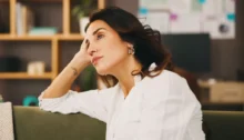 Žena v bílé košili sedí na gauči, opírá hlavu o ruku a zamyšleně hledí do prázdna