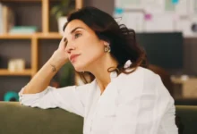 Žena v bílé košili sedí na gauči, opírá hlavu o ruku a zamyšleně hledí do prázdna