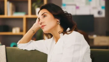 Žena v bílé košili sedí na gauči, opírá hlavu o ruku a zamyšleně hledí do prázdna