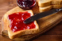Plátek toastového chleba namazaný jahodovým džemem na dřevěném prkénku s nožem