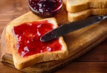 Plátek toastového chleba namazaný jahodovým džemem na dřevěném prkénku s nožem