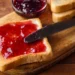 Plátek toastového chleba namazaný jahodovým džemem na dřevěném prkénku s nožem