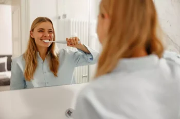žena čistící si zuby elektrickým kartáčkem a její odraz v zrcadle