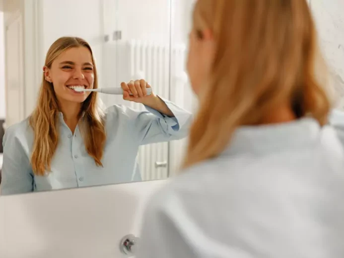 žena čistící si zuby elektrickým kartáčkem a její odraz v zrcadle