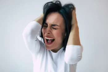 Žena v bílém triku křičí a drží se za hlavu, znázornění emočního vypětí spojovaného s hysterií