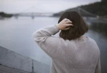Žena stojí u řeky s výhledem na městský most v oblačném počasí, zadní pohled. Osamělá žena za zamračeného dne chodí po městě.
