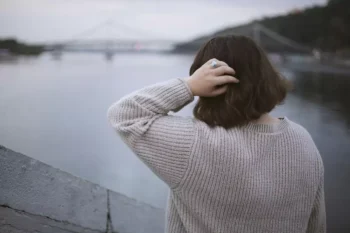 Žena stojí u řeky s výhledem na městský most v oblačném počasí, zadní pohled. Osamělá žena za zamračeného dne chodí po městě.